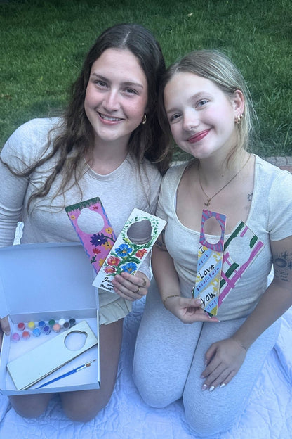 Two young women holding decorated door hanger diy gifts they made outdoors on a girls day outdoor retreat